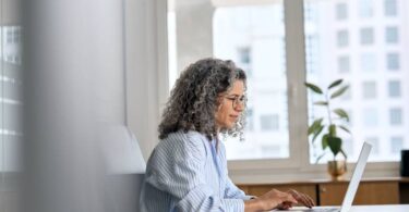Profissional com óculos digitando em laptop ao lado de anotações em ambiente corporativo, simbolizando a avaliação técnica para implementar agentes de IA com suporte especializado.