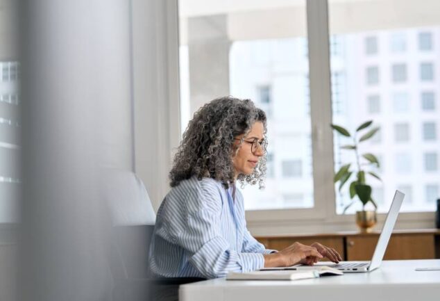 Profissional com óculos digitando em laptop ao lado de anotações em ambiente corporativo, simbolizando a avaliação técnica para implementar agentes de IA com suporte especializado.