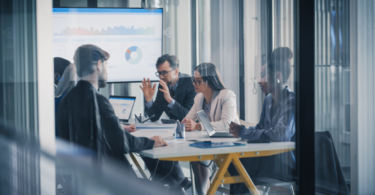 Equipe de líderes comerciais e de dados reunida em sala corporativa moderna, analisando gráficos em uma grande tela e em notebooks para avaliar soluções de prospecção.